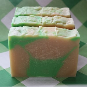 Puede incluir: Tres barras de jabón hecho a mano con un patrón de remolino verde y blanco. El jabón está sobre un fondo a cuadros verde y blanco.
