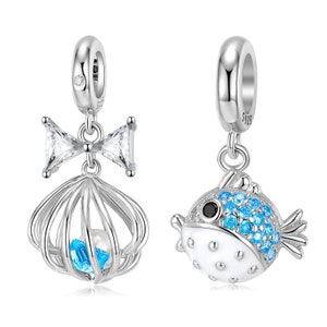 Könnte beinhalten: Zwei silberne Charms. Einer zeigt eine Schleife und eine Muschelkäfig mit einem blauen Edelstein und einer Perle. Der andere Charm ist ein weißer und blauer Kugelfisch mit schwarzen Augen. Beide Charms haben eine Schlaufe.