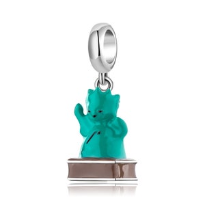 Könnte beinhalten: Ein silberner Anhänger mit einer türkisfarbenen Figur eines Katers, der ein Musikinstrument auf einem braunen Sockel spielt.
