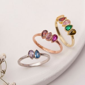 May include: Three gold rings with colourful gemstone accents. The rings feature pear-shaped stones in shades of pink, green, and blue.