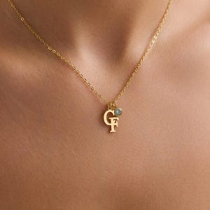 Op de afbeelding: Een goudkleurige ketting met een fijne ketting. De hanger toont de letters "G" en "F" in een gestileerd lettertype, samen met een kleine, ronde, lichtblauwe edelsteen. De ketting wordt om de nek gedragen.