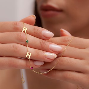 Könnte beinhalten: Eine goldene Kettenkette mit zwei Initialanhängern, einem "R" und einem "H", und zwei kleinen Edelsteinen, einem grünen und einem rosa.