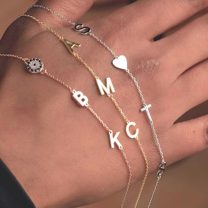 Può includere: Una collezione di delicati braccialetti a catena in oro e argento, ciascuno con un ciondolo unico. I ciondoli includono lettere, un cuore e un design a occhio diabolico. I braccialetti sono mostrati sulla mano di una persona.