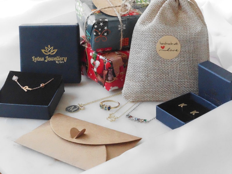 May include: A collection of jewelry in blue boxes, a brown paper envelope, and a burlap bag with a "handmade with love" tag. The jewelry includes a necklace with a charm, a ring, a necklace with a row of colorful stones, and a pair of earrings.