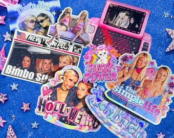 Op de afbeelding: Een verzameling holografische stickers met popcultuurafbeeldingen, waaronder een New York Post krantenknipsel, een roze speelgoedtelefoon, een kat met de tekst PUSSY POWER, en ontwerpen met de teksten HOLLYWEIRD, THE SIMPLE LIFE en ION BITCH.