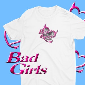 May include: White t-shirt with the text "Bad Girls" in a pink, glossy font. The shirt features a pink heart-shaped graphic with the words "Bad Girls Club" and images of women. The background is light blue.