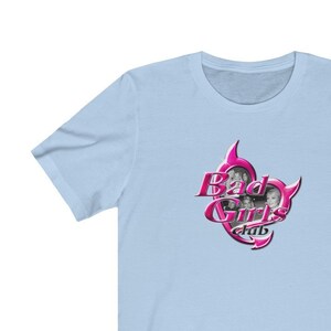 May include: Pale blue t-shirt with a pink and silver graphic. The graphic says "Bad Girls Club" and features images of three women. The text is in a decorative font. The shirt is made of a soft material.