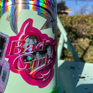 May include: A light green water bottle with a pink and red sticker that says "Bad Girls Club". The sticker has a photo of several women and a pink border. The bottle also has a holographic sticker.