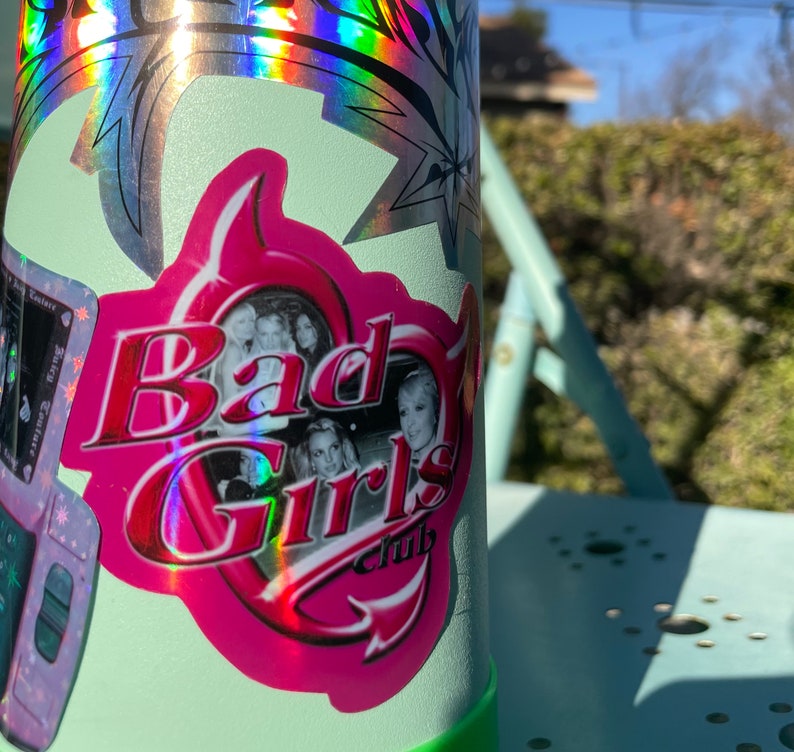May include: A light green water bottle with a pink and red sticker that says "Bad Girls Club". The sticker has a photo of several women and a pink border. The bottle also has a holographic sticker.