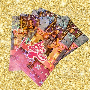 May include: A stack of postcards with a pink and silver design. The postcards feature a woman in a leopard print dress with the text "I Love New York" and star accents. The background is gold.