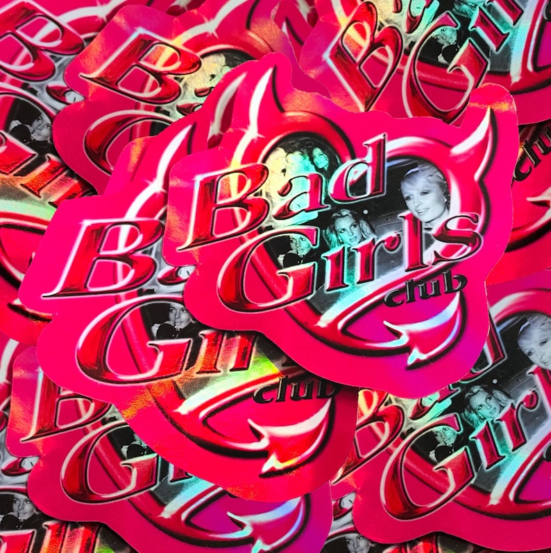 May include: A collection of pink stickers with the text "Bad Girls Club" in a red, glossy font. The stickers feature a devil horn design and images of women. The stickers have a holographic, iridescent finish.