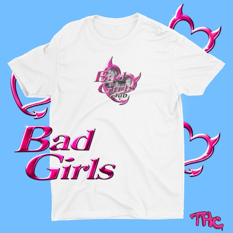 May include: White t-shirt with the text "Bad Girls" in a pink, glossy font. The shirt features a pink heart-shaped graphic with the words "Bad Girls Club" and images of women. The background is light blue.