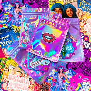 Pu&ograve; includere: Diverse bustine con cerniera olografiche iridescenti con adesivi colorati. Gli adesivi includono un design di labbra rosse, la parola "assy" e le parole "Social Anxiety". Lo sfondo presenta copertine di riviste con testo come "Passport to Paris".
