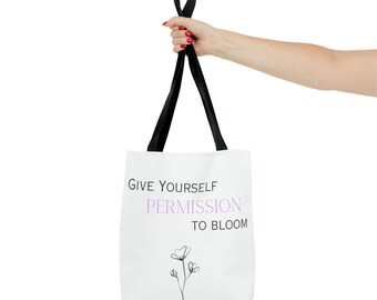 May include: White tote bag with black straps. The bag has the text "Give yourself permission to bloom" printed on it in black with a purple flower graphic.