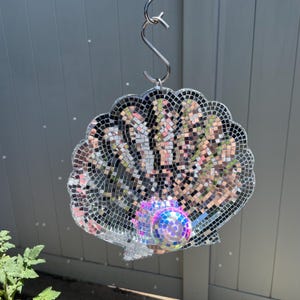 May include: A seashell-shaped hanging ornament covered in small, reflective silver mirror tiles. A single, iridescent, spherical disco ball is attached to the center of the shell. The entire piece is suspended from a silver-colored metal S-hook.