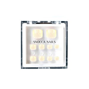 Puede incluir: Un juego de uñas postizas para los dedos de los pies en una caja de plástico transparente. Las uñas son de color amarillo claro con purpurina. El texto "SNOCCO NAILS" está impreso en la caja.