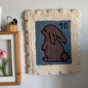 May include: A handmade, textured wall hanging featuring a brown bunny on a blue background, resembling a postage stamp. The bunny is set against a blue backdrop, with the number "10" visible. The border is a cream color with a scalloped edge.