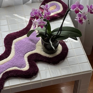May include: A decorative, wavy rug in shades of burgundy, lavender, and cream, with a circular accent piece. A potted orchid with vibrant pink flowers sits on a white tiled table, adding a touch of nature to the scene.
