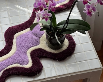 May include: A decorative, wavy rug in shades of burgundy, lavender, and cream, with a circular accent piece. A potted orchid with vibrant pink flowers sits on a white tiled table, adding a touch of nature to the scene.