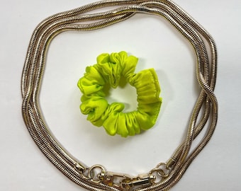 May include: A gold chain necklace with a clasp closure. The chain is made of multiple strands of thin, linked metal. A bright green fabric scrunchie is in the background.