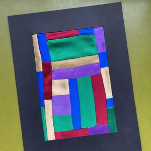 May include: A colorful abstract textile art piece with a patchwork design. The artwork features rectangular shapes in various colors, including green, blue, red, purple, and gold, set against a black background. The piece is framed in a black mat.