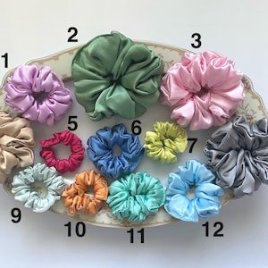 May include: A collection of twelve satin hair scrunchies in various colors, including lavender, sage green, pink, beige, magenta, blue, lime, gray, light blue, orange, teal, and sky blue, arranged on a decorative oval platter with numbered labels.