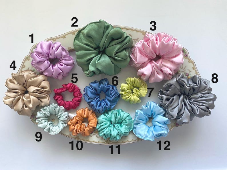 May include: A collection of twelve satin hair scrunchies in various colors, including lavender, sage green, pink, beige, magenta, blue, lime, gray, light blue, orange, teal, and sky blue, arranged on a decorative oval platter with numbered labels.