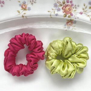 May include: Two circular hair scrunchies with a gathered, pleated fabric texture. One scrunchie is a deep, vibrant magenta pink, and the other is a bright, light lime green. Both items feature a smooth, satin-like finish with a soft sheen.