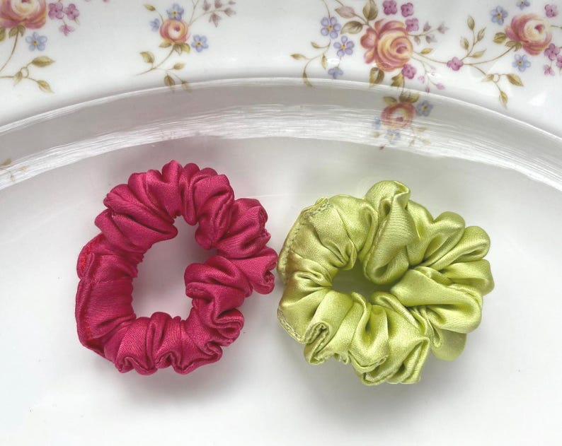 May include: Two circular hair scrunchies with a gathered, pleated fabric texture. One scrunchie is a deep, vibrant magenta pink, and the other is a bright, light lime green. Both items feature a smooth, satin-like finish with a soft sheen.