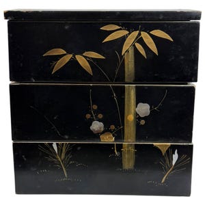 May include: A three-tiered rectangular black lacquer box featuring gold-toned painted bamboo stalks and leaves across the front panels, with small silver and gold floral accents integrated into the design on the middle and bottom sections.