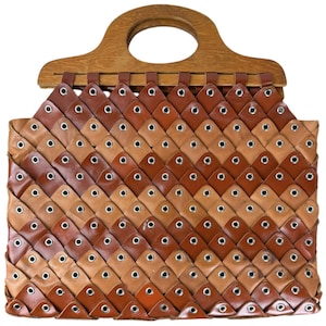 Puede incluir: Bolso de mano de cuero tejido con asa de madera. El bolso presenta un patrón repetitivo de piezas de cuero marrón y beige, cada una asegurada con ojales plateados. El asa es de madera marrón claro con una abertura circular.