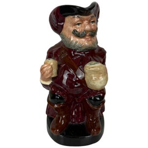 Peut inclure: Une cruche en céramique représentant un personnage vêtu d'un manteau rouge foncé et d'un chapeau noir. Le personnage porte une barbe et une moustache grises, tenant une petite tasse claire dans la main droite et un récipient arrondi plus grand.
