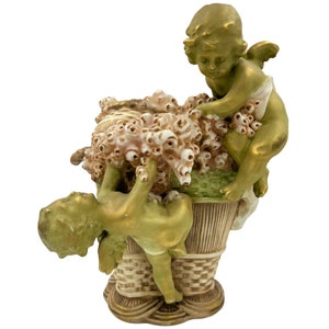 Puede incluir: Una figura decorativa de cerámica que muestra dos querubines dorados con alas interactuando con una cesta grande y texturizada llena de elementos florales blancos y marrones. La cesta tiene un patrón trenzado y una base acampanada en oro y crema.