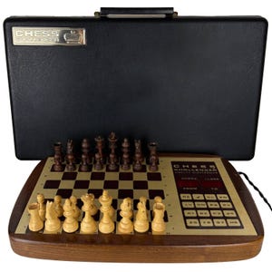 May include: A vintage electronic Chess Challenger game set featuring a wooden board with light and dark squares, a gold-toned control panel with buttons and digital displays, and a set of dark and light wooden chess pieces, all with a black case.