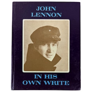 Op de afbeelding: Een hardcover boek getiteld John Lennon In His Own Write met een donkerpaarse kaft en lichtblauwe tekst. Het midden bevat een sepia-kleurige foto van John Lennon met een donkere pet en jas, omlijst door een witte rand op de voorkant.