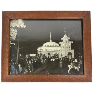 May include: A black and white photograph in a wooden frame shows a building with a sign that reads "CHARIOTEER" and a crowd of people in front of it.