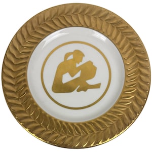 以下が含まれることがあります： 金色の葉模様の縁が付いた丸い装飾用プレート。 プレートの中央は白で、容器を持っている人の金のシルエットがあります。 プレートは装飾用です。