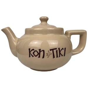 May include: Tan ceramic teapot with a lid and handle. The teapot has a spout and the words "Koh Tiki" in brown lettering on the front. The teapot is on a white background.