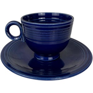 May include: A deep cobalt blue ceramic teacup and matching saucer set. The teacup features a rounded bowl shape with horizontal embossed rings around the exterior and a circular handle. The saucer has a matching concentric ring design.