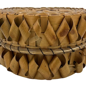 May include: A round, lidded basket crafted from light brown woven wood strips. The exterior features a decorative, looped weave pattern around the sides. A thin, dark-wrapped rim separates the lid from the base, creating a textured appearance.