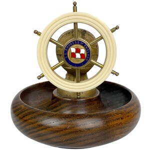 May include: A vintage ashtray featuring a ship's wheel design. The wheel is cream-colored with brass accents and a central emblem. The ashtray has a dark wood base with a circular bowl. The emblem reads "S.S. Empress of England."