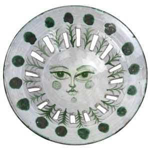 Puede incluir: Un plato decorativo de cerámica con un diseño de cara de sol. El plato presenta una base blanca con detalles en verde, incluyendo una cara de sol en el centro y follaje verde decorativo. El plato tiene cortes rectangulares.