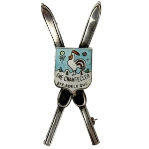 May include: A silver-toned metal brooch shaped like two crossed skis behind a shield-shaped emblem. The emblem features a white rooster, a light blue sky, green trees, and the text THE CHANTECLER STE. ADELE QUE in black lettering.
