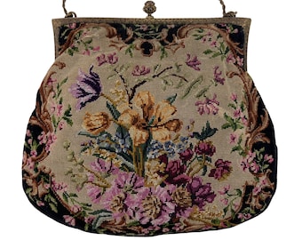 May include: A handbag featuring a needlepoint floral design on a beige background with black borders. The bag has a metal frame with a decorative clasp and a thin metal chain handle. The floral pattern includes yellow, purple, and pink flowers.