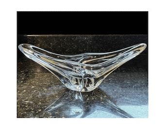 May include: A clear glass bowl with an elongated, boat-like shape and smooth, flowing curves. The vessel features thick, transparent walls that taper toward the pointed ends, creating a fluid, sculptural form with a deep, open center.