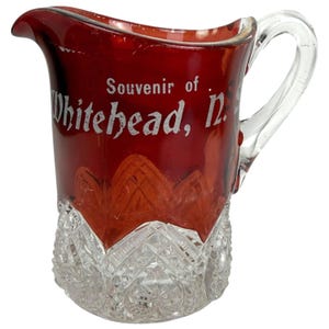 May include: A vintage red glass jug with a clear handle and a cut-glass base. The jug features the words "Souvenir of Whitehead, N." in white lettering. The red glass is semi-transparent, showing the cut-glass design.