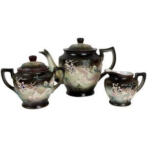 May include: A three-piece tea set featuring a teapot, a lidded sugar bowl, and a creamer. Each piece is crafted from dark brown ceramic with a gradient finish, decorated with a raised, textured dragon motif in white, blue, and green accents.