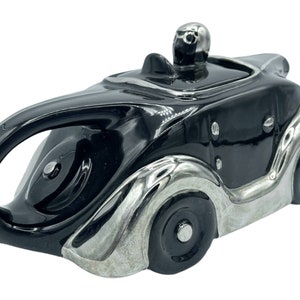 Puede incluir: Tetera de cerámica negra y plateada con forma de coche antiguo. La tetera tiene tapa y pico. El coche tiene detalles cromados y ruedas.