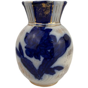 Puede incluir: Un jarrón pequeño, redondo, de cerámica blanca con un diseño floral azul y dorado. El jarrón tiene una boca ancha y un cuello estrecho.