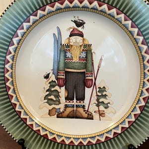 May include: Decorative plate with a winter theme. The plate features a central image of a skier nutcracker figure with skis, poles, and a bird on his head. The border has a green and white striped pattern with a zigzag design.